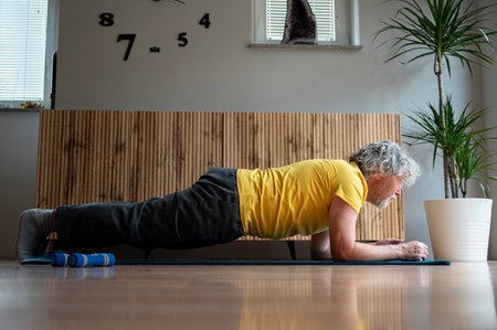 Senior man with grey curly hair in bright yellow tshirt exercising at home making a plank position....