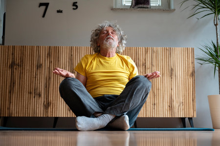Senior man with grey curly hair, wearing a bright yellow tshirt, sitting in lotus position meditating or making a mindfulness practice. At home in a living room on an exercise mat.の写真素材