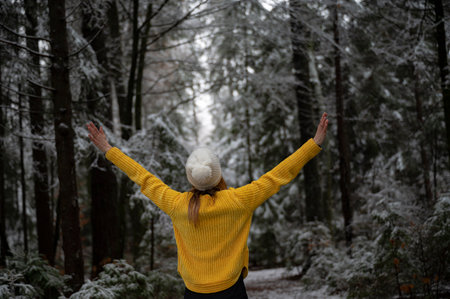 Young woman in bright yellow sweater celebrating life as she stands with her arms raised in the middle of a snow covered winter forest.の写真素材