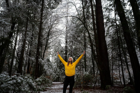 Blissful young woman in bright yellow sweater enjoying herself with arms raised high standing in cold winter forest.の写真素材