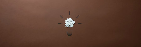 Wide view image of a light bulb made of scattered puzzle pieces and hand drawn shining rays. Over brown background.の写真素材