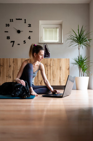 Young woman in casual clothing sitting on a mat in domestic living room using a laptop computer. Her black dog lying next to her.の写真素材
