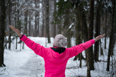 View from behind of a woman in pink sweater standing in calm snow covered winter forest with her arms spread. Enjoying and celebrating life.の写真素材
