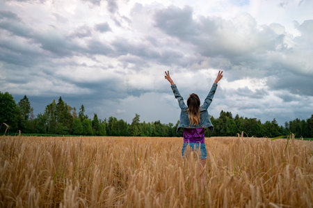 Rear view of a young woman standing in golden summer wheat field enjoying life with her arms raised high in victory.の写真素材