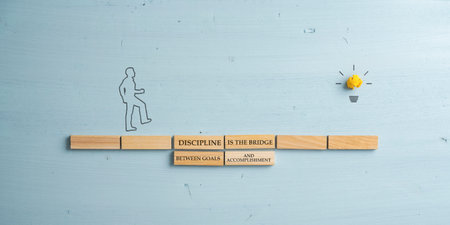 Discipline is a bridge between goals and accomplishment sign written on a bridge made of wooden pegs.. Silhouette of a man walking across it towards a light bulb.の写真素材