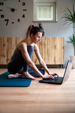 Casual young woman sitting on an exercise mat on a living room floor using a laptop computer to work, study or set up an online class.の写真素材