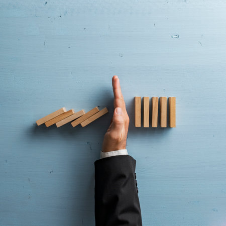 Overhead view of hand in a business suit stopping or intervening collapsing dominos. Over light blue background. Conceptual of crisis management and proactive solution.の写真素材