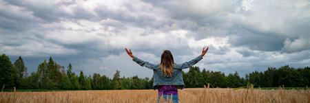 View from behind of a young woman celebrating life or meditating, standing in golden wheat field with her arms open wide towards a cloudy summer sky.の写真素材