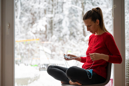 Young woman sitting by the window and knitting using cotton yarn. Wiev of a snowy winetr nature through the window.の写真素材