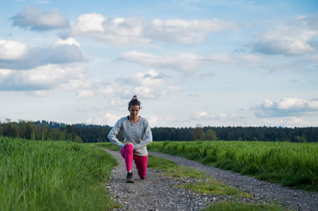 Authentic young woman in pink leggings stretching outside in the middle of beautiful green fields. Warming up before she goes for a run.の写真素材