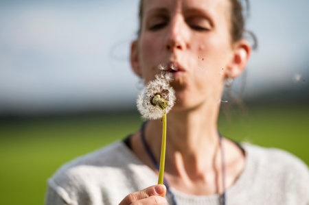 Young woman blowing danelion bulb for the sseds to fly into the air.の写真素材