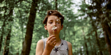 Boy in his early teens blowing dandelion bulb to spread the seeds in a beautiful green spring forest.の写真素材