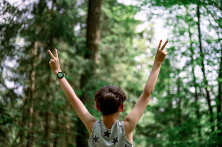 View from behind of a boy in his eary teens standing with his arms raised high in victorious gesture in beautiful green spring forest.の写真素材