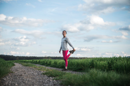 Young woman in pink leggings stretching and warming up before going for a run standing on country road in the middle of beautiful green spring fields.の写真素材