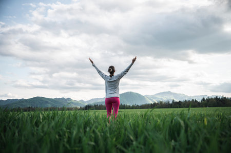 Low angle rear view of a woman standing in the middle of green field or meadow enjoying life and meditating with her arms raised high. Under cloudy spring sky.の写真素材