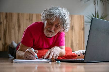 Senior man lying on living room floor taking notes in his notebook as he attends an online class using laptop computer.の写真素材