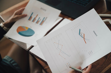 Over the shoulder view of a woman reviewing financial business documents with graphs and charts in a casual domestic workspace.の写真素材
