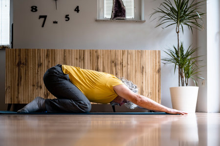 Profile view of a senior man stretching in a child pose exercise on a mat in domestic living room. Conceptual image of active retirement and care for ones health.の写真素材