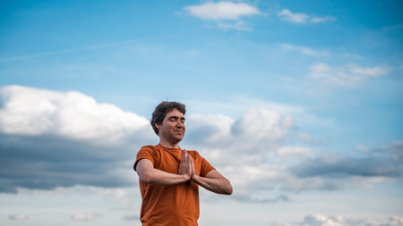 Young man praying or meditating with his palms joined in front of his chest. Standing under beautiful blue sky with white clouds.の写真素材