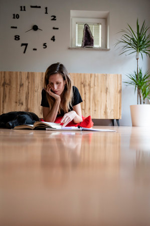 Low angle view of a young woman, a student, lying on a living room floor reading a textbook and...