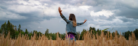 Wide view image of a young woman celebrating and enjoying life standing with her arms spread widely in the middle of golden wheat field under a dramatic cloudy summer sky.の写真素材