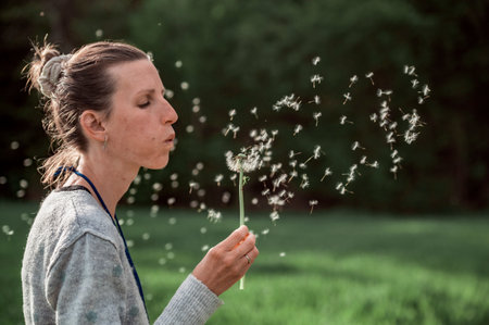 Young woman blowing a delicate dandelion flower bulb with seeds floating all around her.
