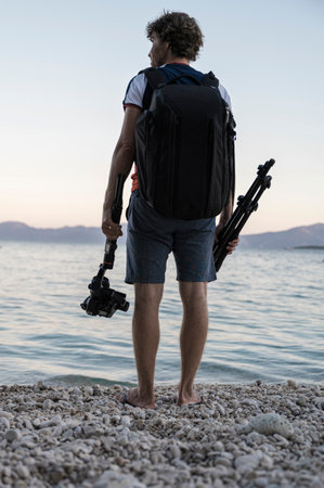 View from behind of a young professional photographer with a tripod and gimbal camera holder in his hands standing on a beautiful pebble beach by a calm sea.の写真素材