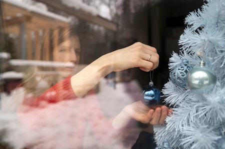View through a window of a young woman decorating a white christmas tree hanging a blue holiday bauble on it. Snowy nature reflecting in the glass.の写真素材