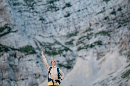 Happy young female hiker with one arm raised in victory. Standing with high rocky mountains in background.の写真素材