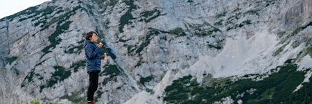 Profile view of a boy standing on mountain peak with high rock mountain in background, blowing soap bubbles. Wide view image.の写真素材