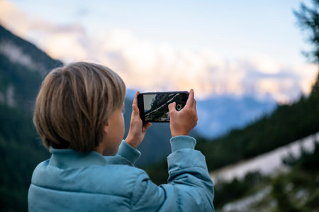 Over the shoulder view of a toddler boy taking a picture with his smart phone of beautiful mountains...