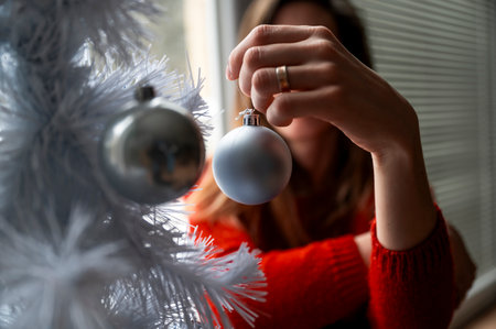 Closeup view of a woman in red sweater hangin shiny blue holiday bauble on a white christmas tree.の写真素材