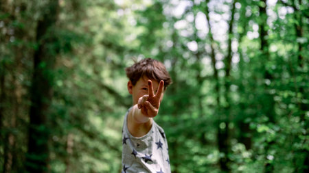 Young teenage boy showing a victory gesture straight towards the camera standing outside in beautiful green nature.の写真素材