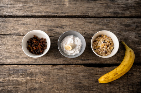 Top view of three bowls with healthy breakfast ingredients - raisins, yoghurt and granola with a ripe banana beside them. Over textured rustic wooden boards.の写真素材