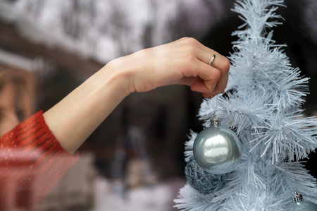 View through the window of a woman hanging shiny blue holiday bauble on a white christmas tree. With snowy winter nature reflecting in in the glass.の写真素材