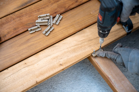Overhead closeup view of worker hands in protective gloves screwing a metal screw in newly layed wooden plank for outdoor patio with an electric machine.の写真素材