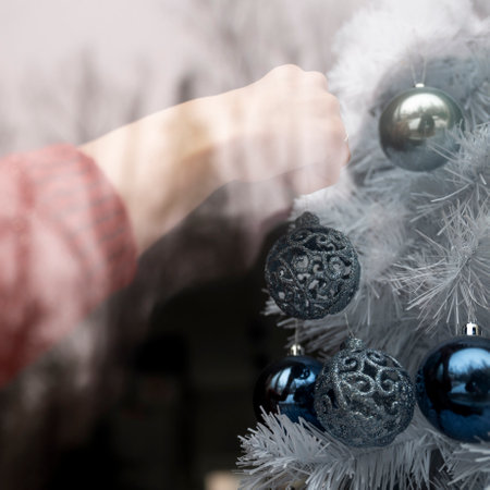 View through a window of female hands decorating white christmas tree with shiny blue holiday baubles in a seasonal image of jolly new year holidays. With reflection of the outside in the glass.の写真素材