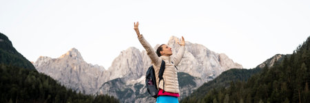 A woman stands triumphantly with her arms raised towards the sky, surrounded by towering mountain peaks and lush greenery. Dressed in a light jacket and carrying a backpack, she appears to be enjoying a moment of exhilaration and freedom amidst the majestic mountain landscape. The scene captures a sense of adventure, serenity, and connection with nature.の写真素材