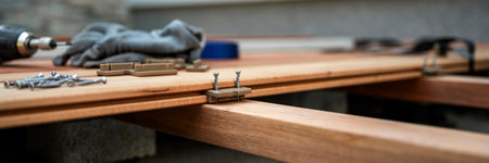 A detailed view of the deck installation process, focusing on hidden fasteners and screws being used to secure wooden deck boards. A power drill, screws, and fastening clips are visible, along with gloves in the background. The setup highlights the precision of aligning and securing deck boards without visible screws on the surface.の写真素材