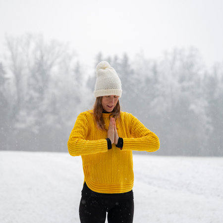 Spiritual young woman with a hat and bright yellow sweater standing in the middle of peaceful cold winter nature praying or practicing gratitude and mindfulness with her palms joined in front of her chest.の写真素材