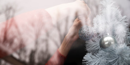 The image captures a serene moment of decorating a white Christmas tree. A hand gently places a blue ornament adorned with intricate patterns onto the tree, adding to the festive ambiance. The soft focus and delicate lighting create a dreamy, peaceful holiday atmosphere with a view through a window.の写真素材