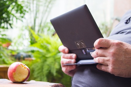Male hands using tablet computer and bitten apple on wooden table. Winter garden background.の写真素材