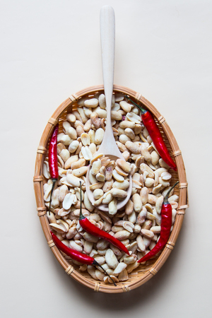 Peeled salted peanut with red hot chili pepper in wicker basket and wooden spoon on beige backgroundの写真素材