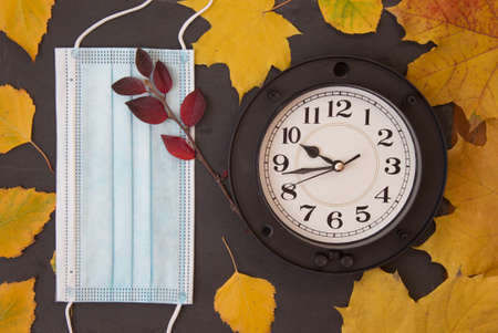 Blue face medical mask, wall clock and colorful autumn leaves. Top view.