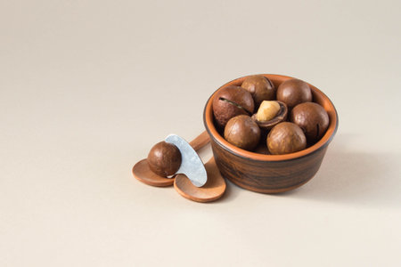 Macadamia nuts in ceramic bowl on beige background. Nuts with sawn nutshells and with opener key on the wooden spoon. Healthy snack, superfood.の写真素材