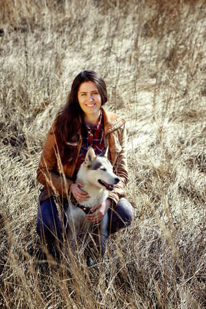 Girl sitting in field with husky and smiles . Open look. Beautiful young brunette with long hair,...