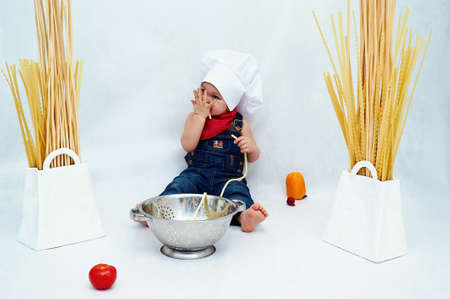 Little boy sitting barefoot in a denim jumpsuit , the colander with the pasta and eatの写真素材