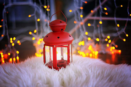 Red candle holder with a candle stands on white skin. Bokeh garland.A warm Christmas atmosphereの写真素材
