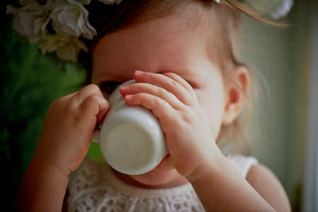 Baby in wreath and dress with tulle and lace sits on the windowsill, and drink of that Cup. Tenderness and feeling of lightnessの写真素材