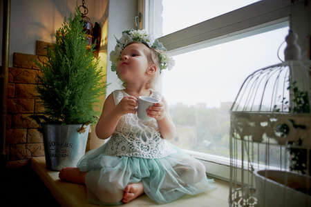 Baby in wreath and dress with tulle and lace sits on the windowsill, and drink of that Cup. Tenderness and feeling of lightnessの写真素材